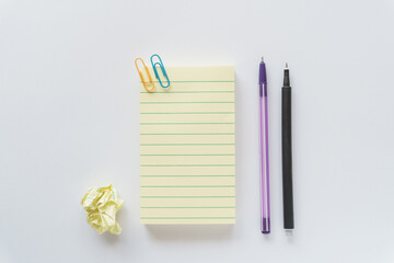 Top-down horizontal flat lay of a blank yellow lined note pad with paper clips, pens, and a crumpled yellow paper ball on a white surface, representing brainstorming and trial and error. © Karina Dolzhenko