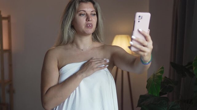Young woman in a bedsheet flirts online while using a smartphone at home, representing webcam modeling, private communication and dating.