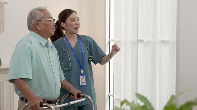 Female Asian caregiver walks beside senior Asian man with walker, pointing ahead during gentle mobility training in assisted living center, encouraging confidence, rehabilitation and active aging.