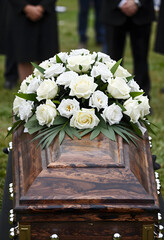 Obraz premium Funeral service and bereavement, white rose floral arrangement on a wooden casket during a burial ceremony with mourners standing in the background