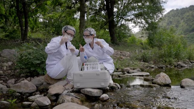 Two professional researchers in protective gear test water samples on a rocky riverbank in a natural outdoor setting.