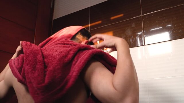 Rear view of an Asian young man with wet bare back taking a red towel and drying his hair in a sunlit bathroom, morning hygiene routine concept