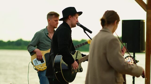 Outdoor riverside band performance with acoustic guitar singer, bass player and audience enjoying live music under wooden canopy during sunset gathering