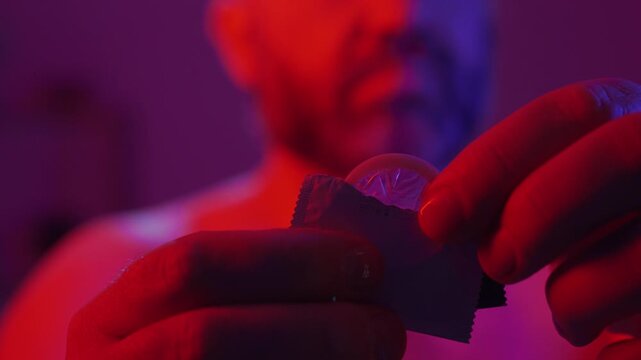 Bearded man holding condom in hand standing in red and blue light