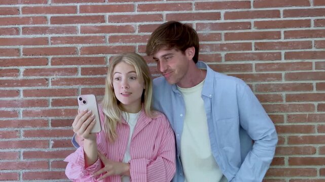 Cheerful young couple standing against a brick wall, smiling and waving during a video call on a smartphone, enjoying a moment of connection with family or friends while communicating online
