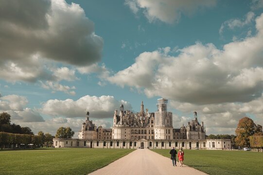 Historic architecture blends with expansive landscape, inviting exploration while walking along the path on a sunny day under a sky filled with clouds near a grand castle