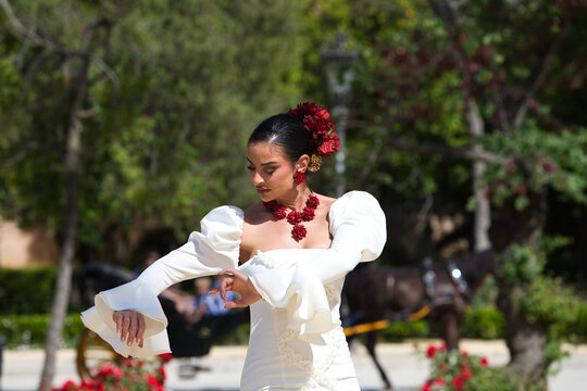 A beautiful woman dancing flamenco in a park in Seville, Spain. She is wearing a traditional white dress with ruffles and has red flowers in her hair. Flamenco, a UNESCO Intangible Cultural Heritage.