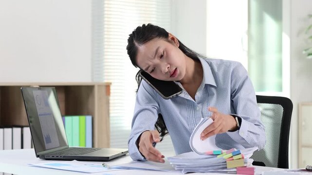Stressed asian businesswoman multitasking at her desk, talking on the phone and sorting through documents, feeling overwhelmed and busy with a heavy workload in the office