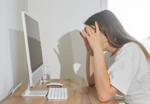 Frustrated businesswoman holding head in hands while sitting in front of computer monitor. Overworked remote employee facing serious system errors and critical project failures. Depressed woman at