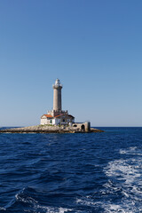 Lighthouse complex stands on a rocky islet as a boat passes across open sea with copy space above. Concept of maritime navigation, vessel travel, coastal landmarks and marine safety. © Natallia