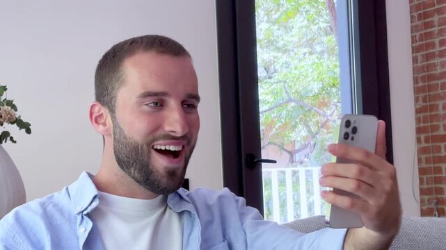 Handsome young man with a beard happily video calling with friends or family on his smartphone, smiling, waving, and gesturing while relaxing on the couch in his living room