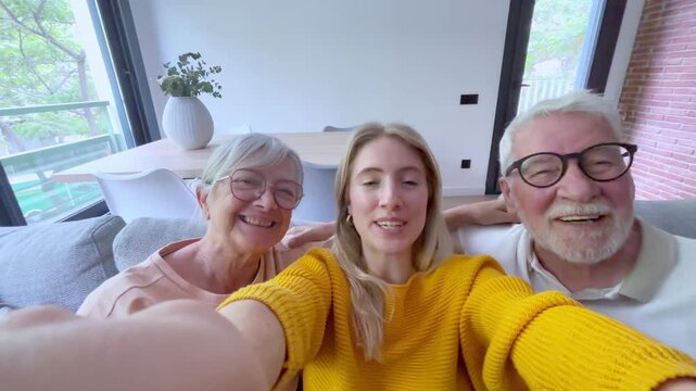 Pov shot of a happy family, featuring a young daughter with her elderly parents, enjoying a video call together, smiling and waving at the camera while sitting on the sofa in their living room