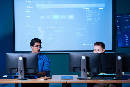Two software developers working on code programming and system engineering in dark office room with computer monitors screen showing workflow interface data