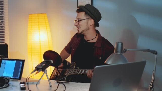 An adult male musician wearing glasses and a fedora plays acoustic guitar and sings into a microphone in a home recording studio with laptops and recording gear.