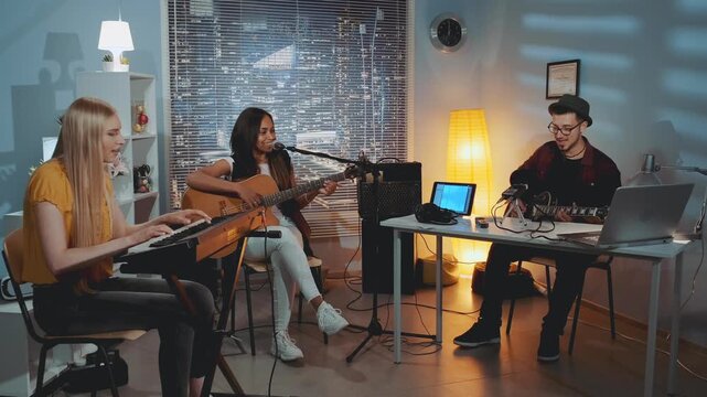 Four young adults play instruments and sing together in a professional indoor recording studio with city views through large windows.