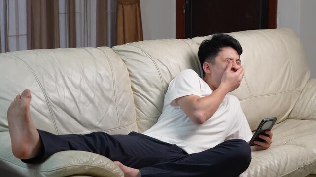 Asian young man lying on a couch holding a smartphone and yawning, tired and lazy weekend concept, horizontal video