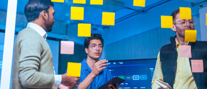 A diverse team of software developers in a modern office brainstorm and plan a project using sticky notes on a glass wall at night.