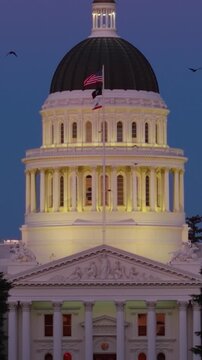 Aerial view shows California State Capitol in Sacramento at night with the dome lit under evening skies and trees nearby.