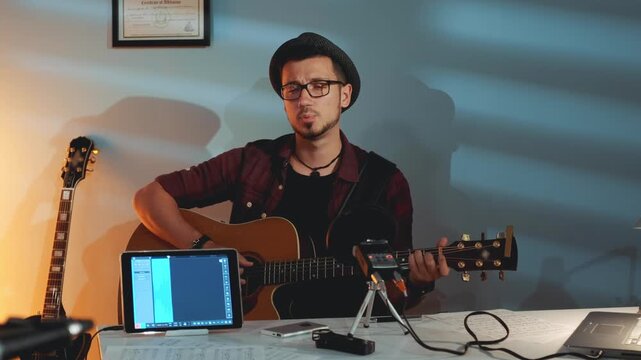 A young man wearing glasses and a fedora plays acoustic guitar while seated at a desk in a home recording studio with multiple guitars and digital equipment.