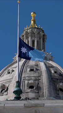Flag is waving atop the State Capitol building in St Paul, Minnesota, showing the state symbol under a blue sky.