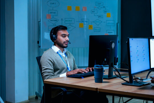 Indian customer support agent wearing headset working on computer late at night in modern dark office setting