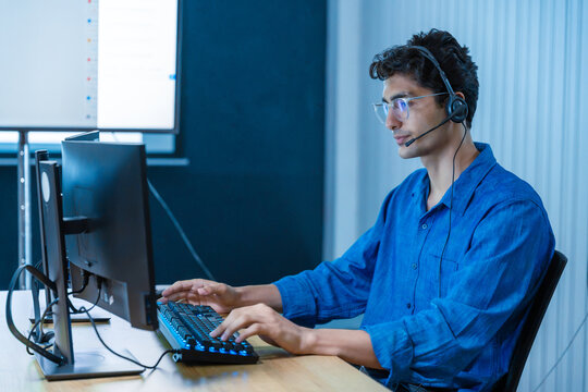Professional young man working as call center representative using dual monitors and headset in contemporary workspace.