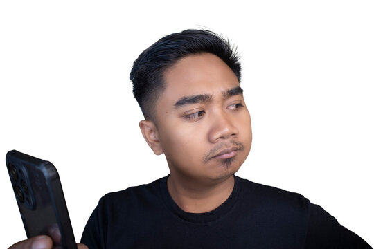 Skeptical Indonesian man wearing a black t-shirt looking away from his mobile phone with a pouting and doubtful expression against a white background.
