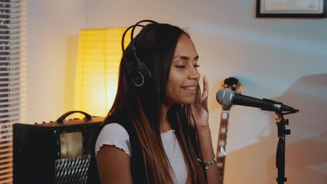 Black female vocalist wearing headphones singing into microphone in a warm, indoor home recording studio at nighttime, smiling and performing with musical equipment around her.