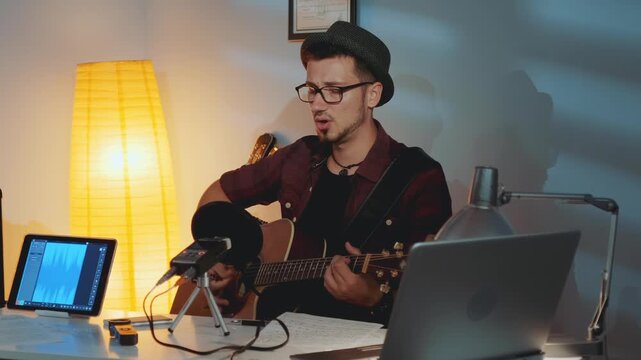 An adult male wearing glasses and a fedora plays acoustic guitar and sings into a microphone in a cluttered home recording studio filled with equipment and warm lighting.