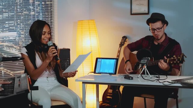 Three young adults rehearse music chords in a professional studio at nighttime. A female vocalist reviews sheet music while a guitarist plays acoustic guitar beside her.