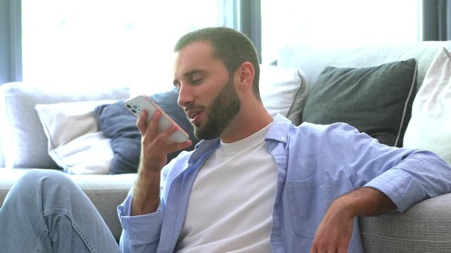 Casual man sitting on the floor in his living room, leaning against the sofa while talking on his smartphone using the speakerphone function, sending a voice message or using an assistant