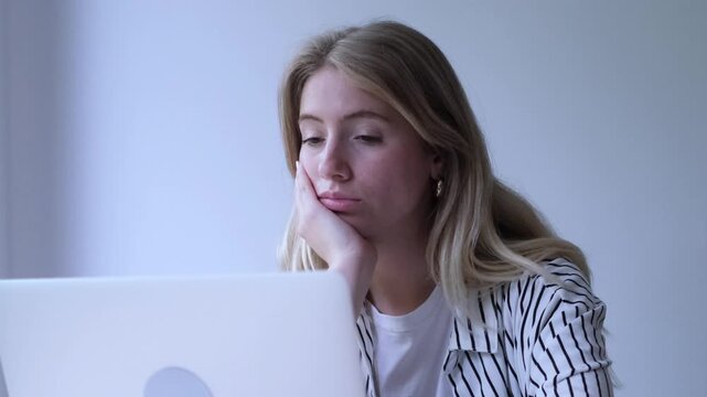 Bored young businesswoman sitting at her desk working on a laptop computer suddenly becomes surprised and happy, smiling and covering her mouth with her hand in disbelief at the good news