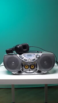 Vertical video silver retro boombox with visible cassette tape and headphones resting on top, placed on a white table against a teal background, evoking vintage music nostalgia and analog sound