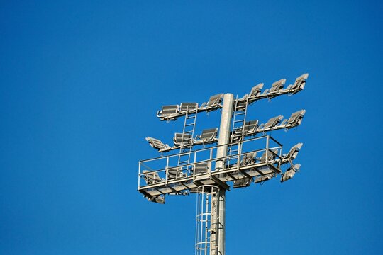 Tall sports lighting tower with multiple floodlights against clear blue sky. The structure is metallic and designed for illuminating outdoor sports events.
