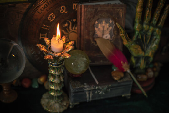 Burning candle in vintage candlestick on mystical altar with crystal ball, zodiac wheel, old spell books and dip pen 
