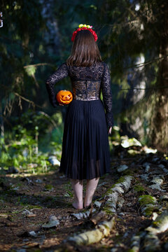 Female stands stands with her back to the camera in the forest with a Halloween pumpkin in her hand.
