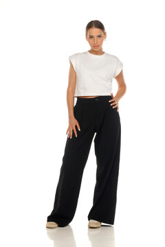 Full length fashion portrait of woman wearing white crop top and black trousers, standing pose, studio shot.