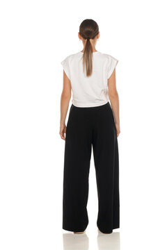 Full length rear view fashion portrait of woman standing, white crop top and black trousers, on white background