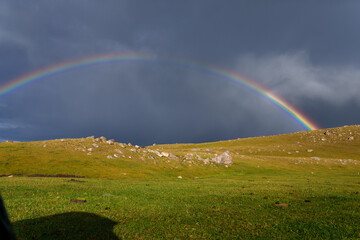 Vivid rainbow arcs across a dark, stormy sky. Green grassy hills stretch beneath the colorful arc. Scene suggests hope after rain, nature's renewal. Perfect for themes like resilience © Pauli