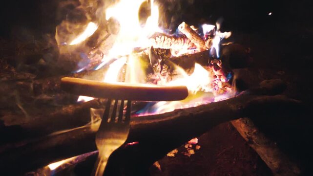 Roasting a sausage on a fork over bright glowing campfire flames at night