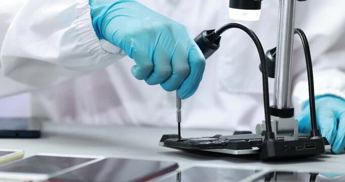 Close-up of a technician in blue gloves using a screwdriver to repair a smartphone at a laboratory workbench with a digital microscope.
