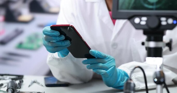 Forensic technician in blue gloves examines a black smartphone beside a microscope in a laboratory, digital evidence analysis and device inspection.