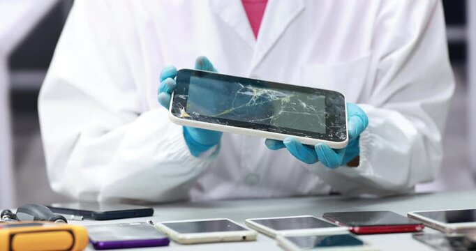 Technician in a white lab coat and blue gloves holds a cracked tablet above a table with damaged smart phones, electronic device repair and screen replacement.