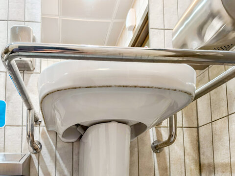 Ceramic sink with a safety grab bar in a public restroom designed for people with limited mobility. Uncleaned view from below. A grab bar for people with disabilities is installed next to the sink