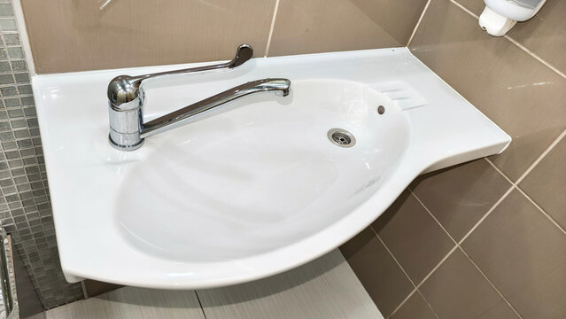 White ceramic corner washbasin with ergonomic chrome faucet in a public bathroom. Handicapped accessible sink installation with modern plumbing design for elderly or disabled