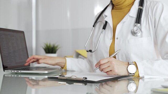 Doctor working on medical records, writing prescription in clinic office, using laptop for patient data management. Medicine, healthcare and science concept