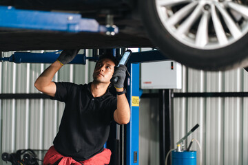 Fototapeta premium Mechanic inspecting vehicle undercarriage on hydraulic lift in automotive repair garage with focused expression