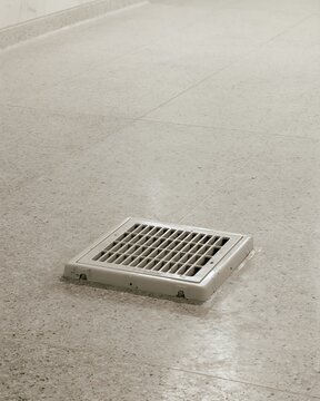 A square metal floor drain grate installed in a light gray speckled terrazzo floor surface, viewed from a angle in a bright room