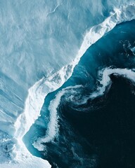 Aerial view of a massive glacier edge meeting deep blue ocean water, showing white ice, cracks, and swirling patterns in the freezing cold sea