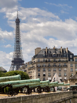 Rows of weathered bronze cannons stand guard at Les Invalides, with the Eiffel Tower rising above Parisian rooftops in the distance.
